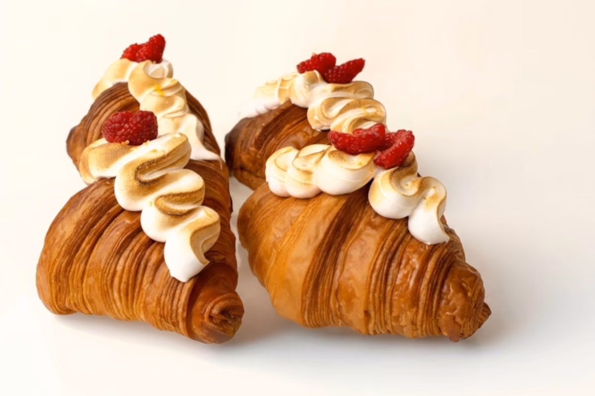 due croissant dorati guarniti con meringa fiammeggiata e lamponi freschi su fondo bianco