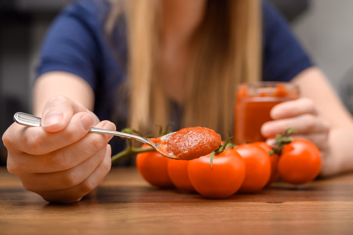 una mano tiene un cucchiaio colmo di concentrato di pomodoro davanti a pomodori freschi