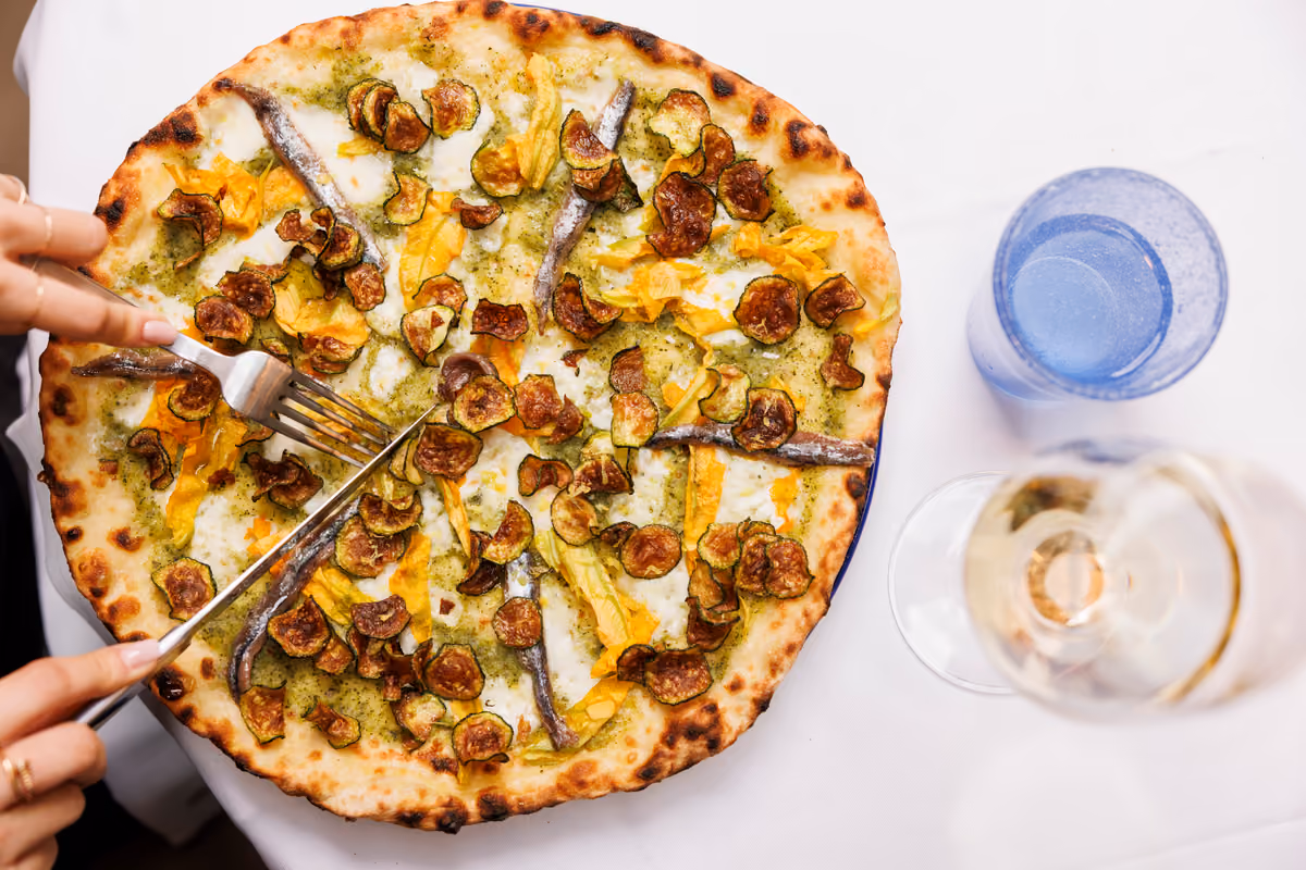 mani che tagliano una pizza gourmet con fiori di zucca, zucchine e acciughe