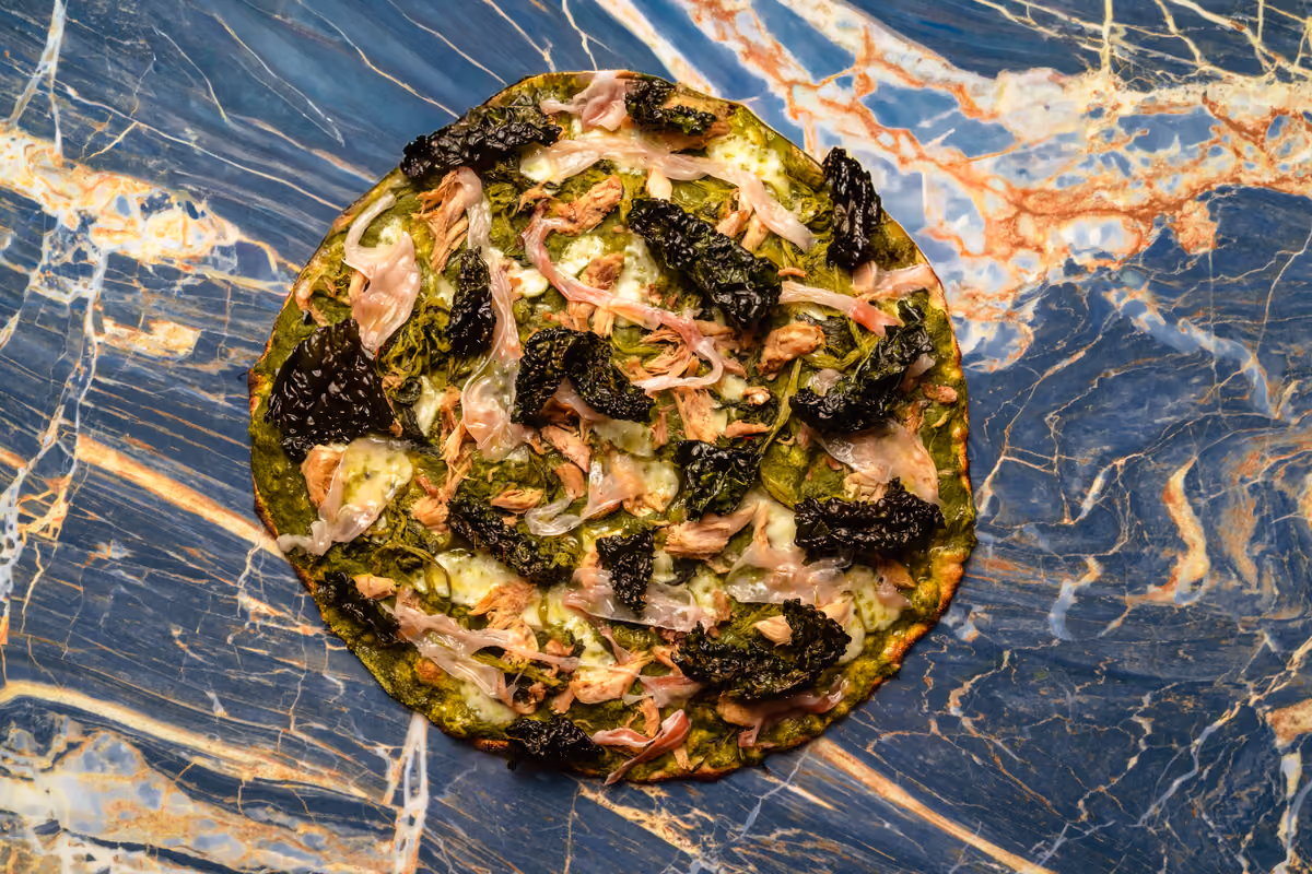 Pizza romana con pesto scuro, verdure croccanti e prosciutto su piano marmoreo blu