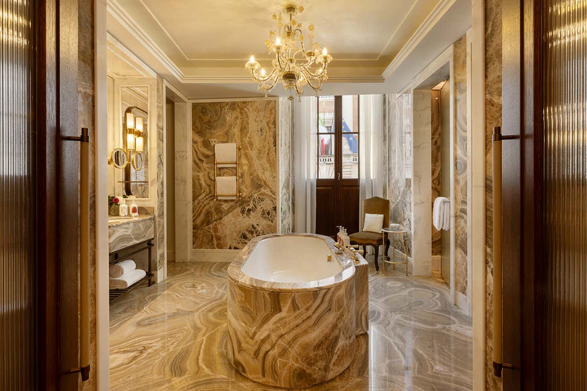 Il bagno padronale della Theodoli Heritage Suite, caratterizzato da un rivestimento integrale in pregiato marmo italiano venato, vasca freestanding centrale e un lampadario artistico in vetro