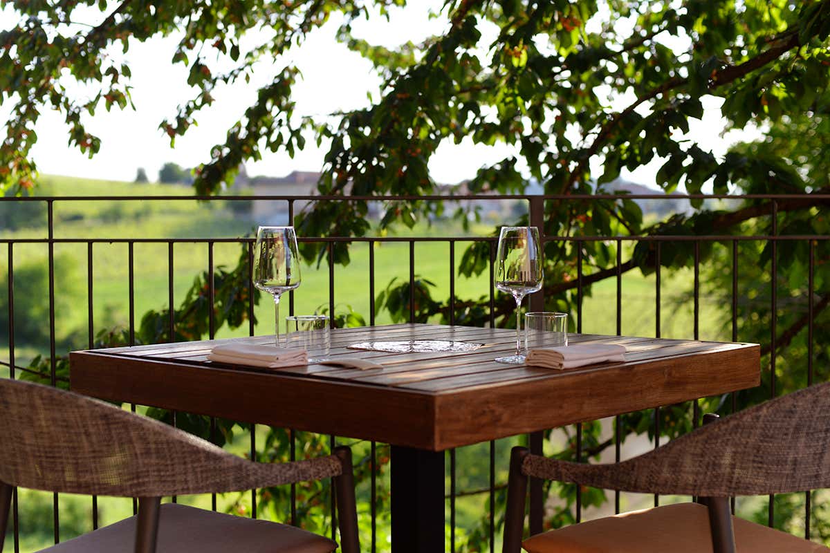 Didascalia: Un tavolo allestito sulla terrazza panoramica di Radici Ristorante in Vigna, incorniciato dal verde delle colline Unesco