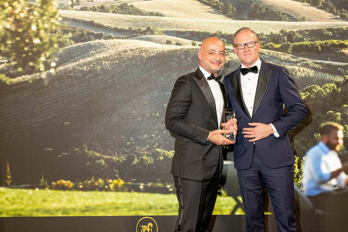 Roberto Protezione (a sinistra), General Manager di Castelfalfi, riceve il premio Best Destination Estate durante la cerimonia. Lo sfondo raffigura le iconiche colline toscane, simbolo della tenuta Castelfalfi